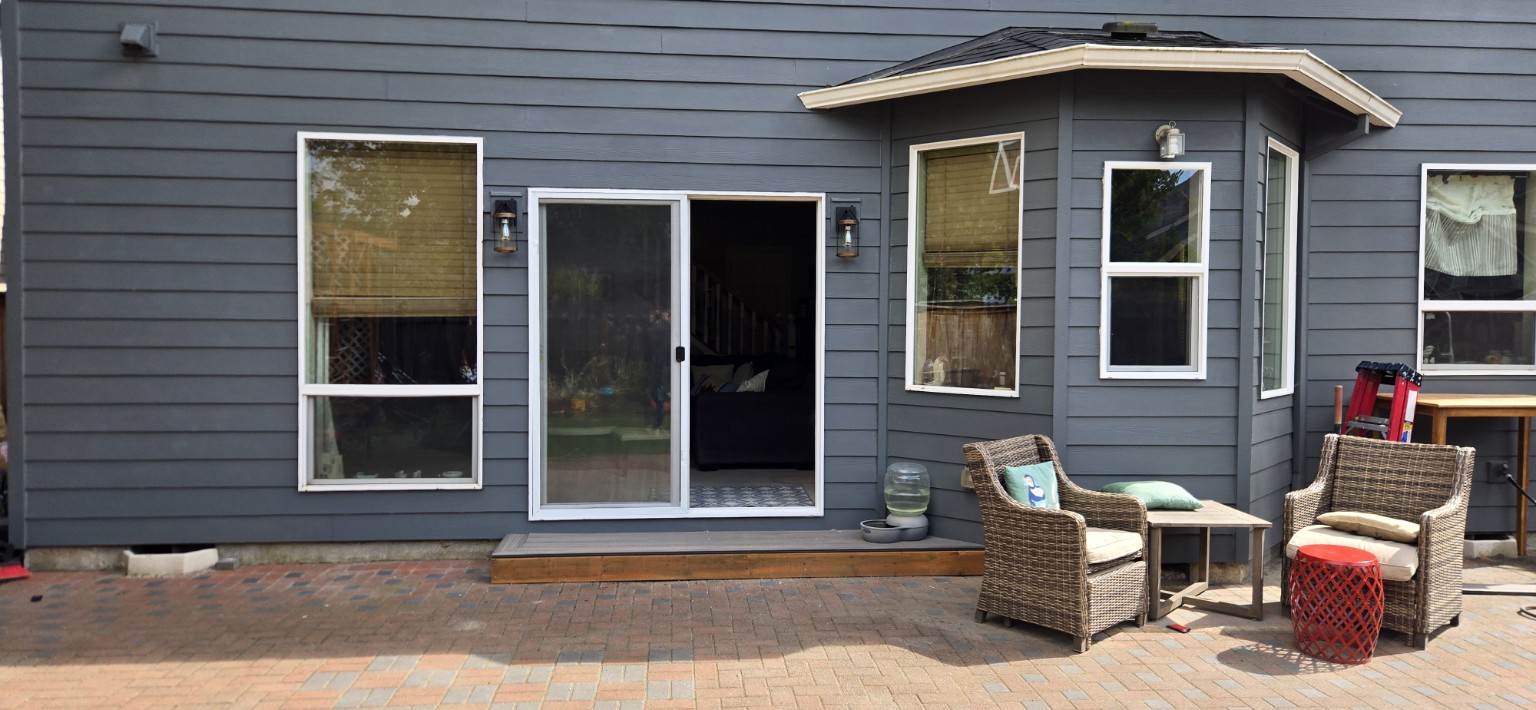 Sliding patio door and white-trimmed windows on a gray home, with a wood step and brick patio